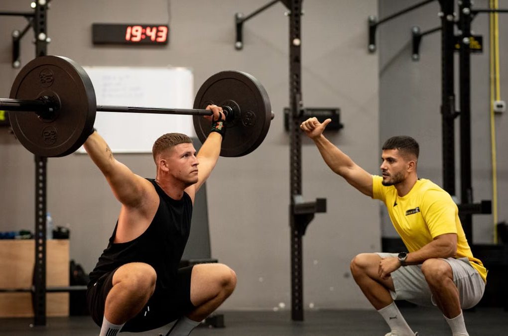 Man lifting weights under coach supervision in a gym setting, focusing on strength training.