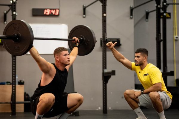 Man lifting weights under coach supervision in a gym setting, focusing on strength training.
